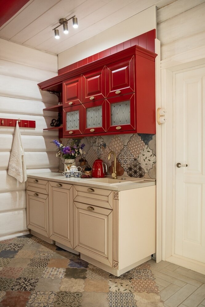 Плитку на фартук и на пол в кухне выбрали одинаковую, чтобы выделить пространство небольшой кухни и отделить ее визуально от гостиной. Плитка в стиле пэчворк с матовым эффектом хорошо сочетается с русским стилем. Ярко-красная кухня с буфетом гармонично вписалась в проект, несмотря на то, что это готовая, а не заказная мебель.
