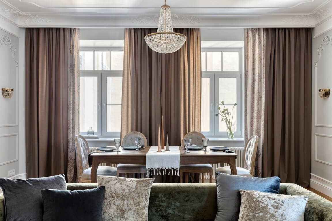 Выраженная текстура зеленого дивана делает пространство «живым», зеленый оттенок воспринимается мягко и уютно. Стол и стулья, расположенные за диваном у светлых окон, являются обеденной зоной. Деревянный элегантный и выразительный стол выстраивает композицию комнаты.