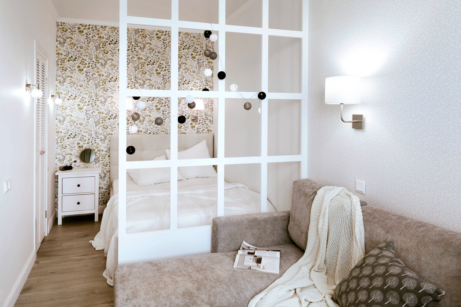 Зонирование в жилой комнате сделали при помощи перегородки из МДФ. Таким образом ограничили приватную зону — спальню. В изголовье кровати поклеили скандинавские обои с цветочным орнаментом — получилось очень уютно.
