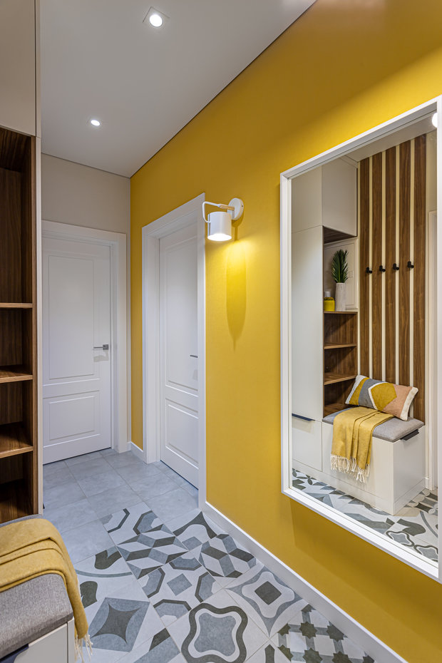 В прихожей выложили нескользкую плитку с интересным геометрическим рисунком, который плавно переходит в фоновую однотонную плитку. На стенах обои, чтобы удешевить стоимость проекта на подготовительных работах, и функциональные рейки, между ними расположены крючки для верхней одежды.