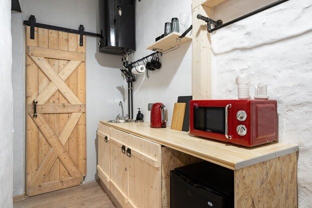 СВЧ-печь яркого контрастного красного цвета специально оставлена в зоне видимости, ее визуально поддерживает красный яркий электрический чайник. Плита спрятана внутрь шкафа. В открытой правой части кухонного шкафа располагается небольшой холодильник.