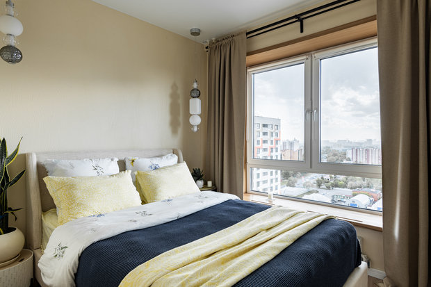 В спальне повторили прием с окном с деревянной облицовкой, на окна повесили шторы-блэкаут. На стенах такие же обои с волнообразным рисунком, как и в кухне-гостиной, только солнечного песочного цвета.