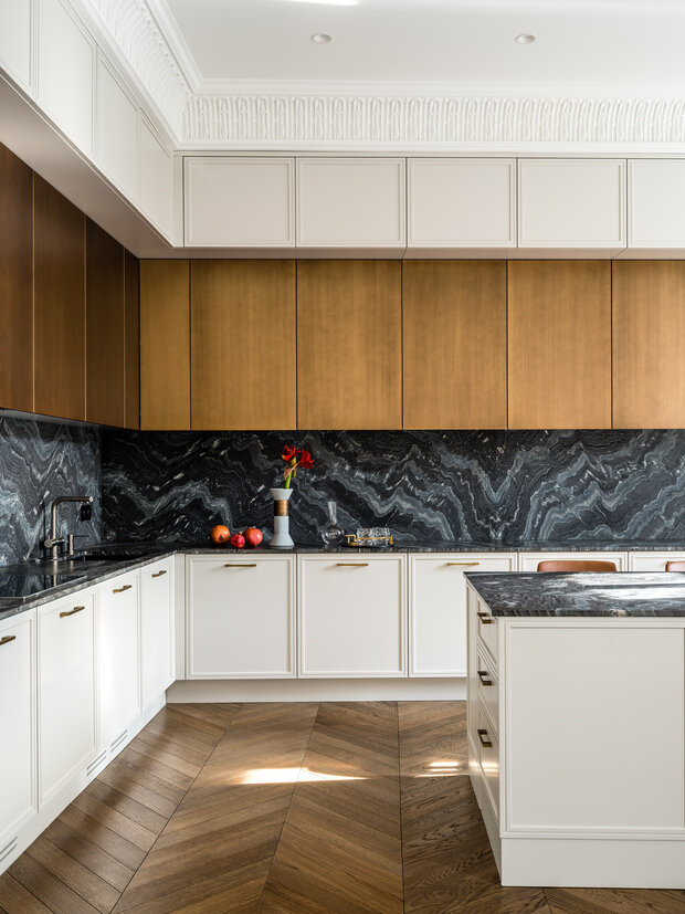 В кухне есть все необходимое: встроенные холодильник, варочная поверхность, духовой шкаф, СВЧ, вытяжка, посудомоечная машина, винный шкаф. Кухню изготовили из МДФ, частично выкрасили фасады эмалью в цвет стен и сделали акцент в виде второй линии фасадов, патинированных под бронзу. Фартук и столешница из матового итальянского гранита с необычной фактурой. Чтобы получить такое красивое перетекание рисунка на камне, пришлось рассмотреть не один вариант раскроя слэбов.