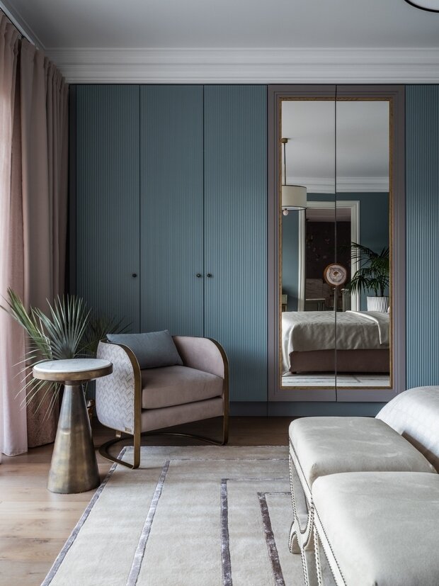 В хозяйской спальне, помимо гардеробной, есть вместительный шкаф во всю стену, фасады сделали рельефными и покрасили в цвет стен. Чтобы он не был монотонным, в центре дизайнер разбавила его зеркальными фасадами в двухцветной раме. Ручки подобрали из розового агата — на зеркале два полукруга как акцент, на остальных фасадах — маленькие кнопки.