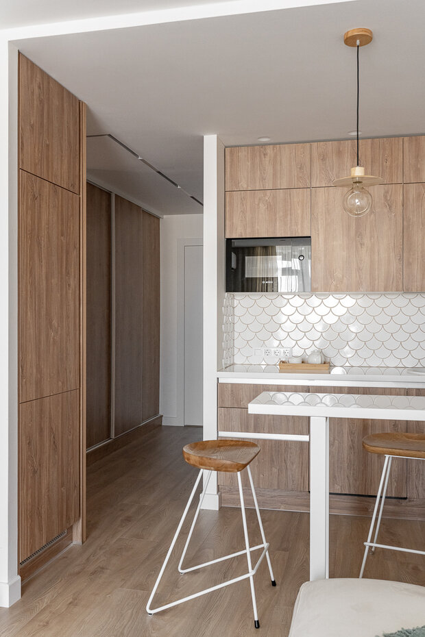 Барная стойка является продолжением рабочей поверхности и своеобразным разделителем кухонной зоны от гостиной. Граница столешницы завязана с объемом холодильника. Колонна с холодильником выдвигается в коридор, ее «подхватили» ритмом из реек и стеллажом. Стеллаж в коридоре, полки выполнены из шпона дуба. Столешница из искусственного камня.