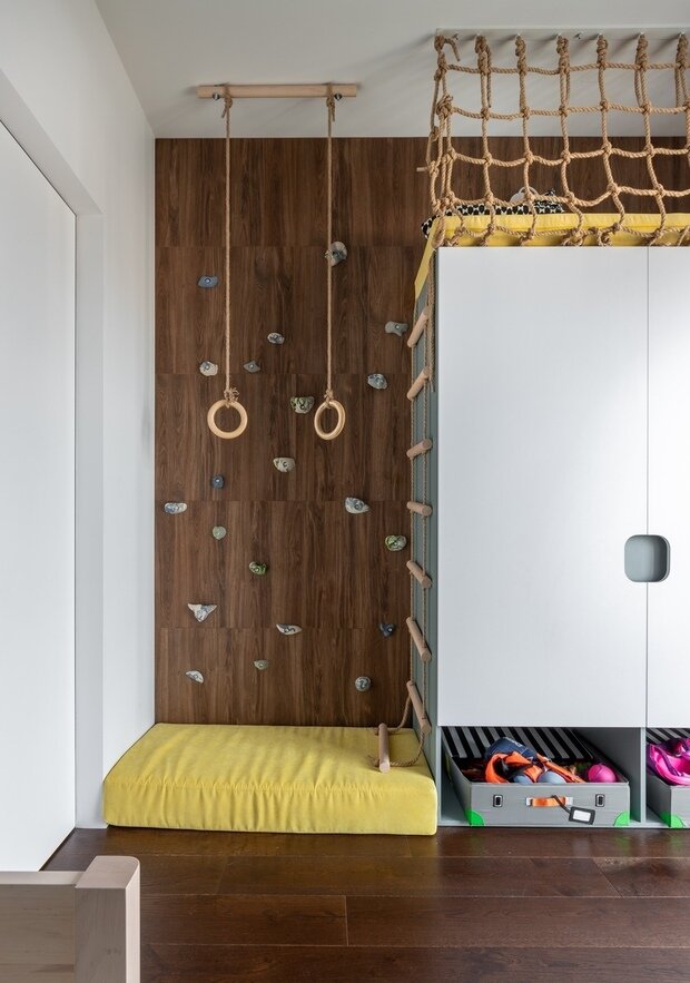 В комнате для мальчика воплотили в жизнь мечту, наверное, каждого ребенка, разместив в игровой зоне скалодром, гимнастические кольца и укромное место на шкафу, в которое можно забраться по веревочной лестнице.