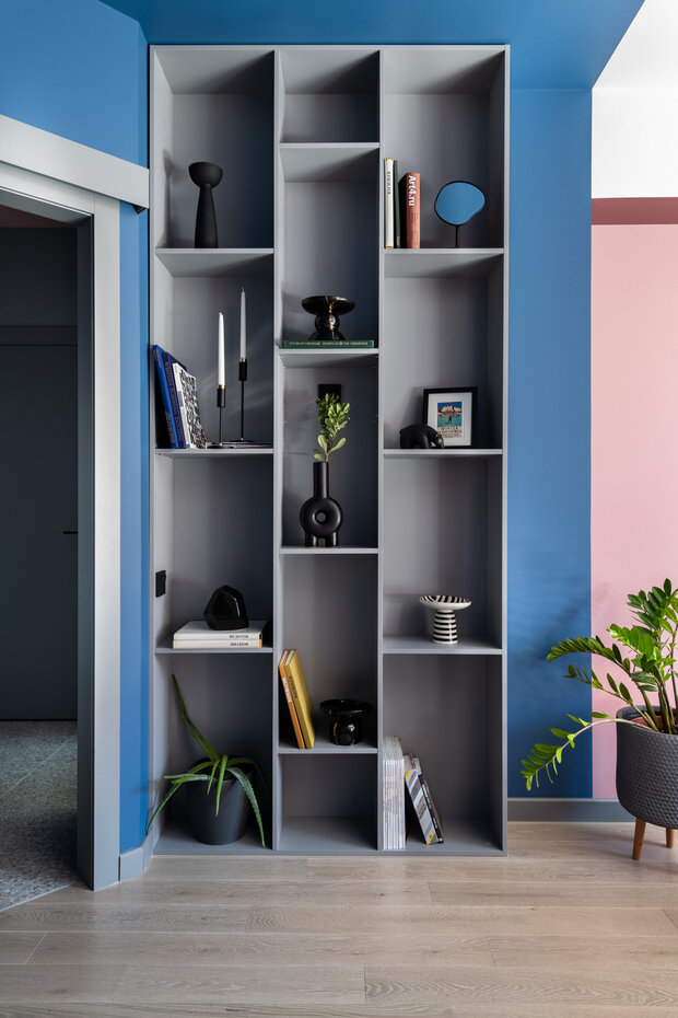 Серый открытый стеллаж выполняет несколько важных функций: он скрывает узкий выступ стены у входа и визуально делит помещение на кофейную зону и спальню.