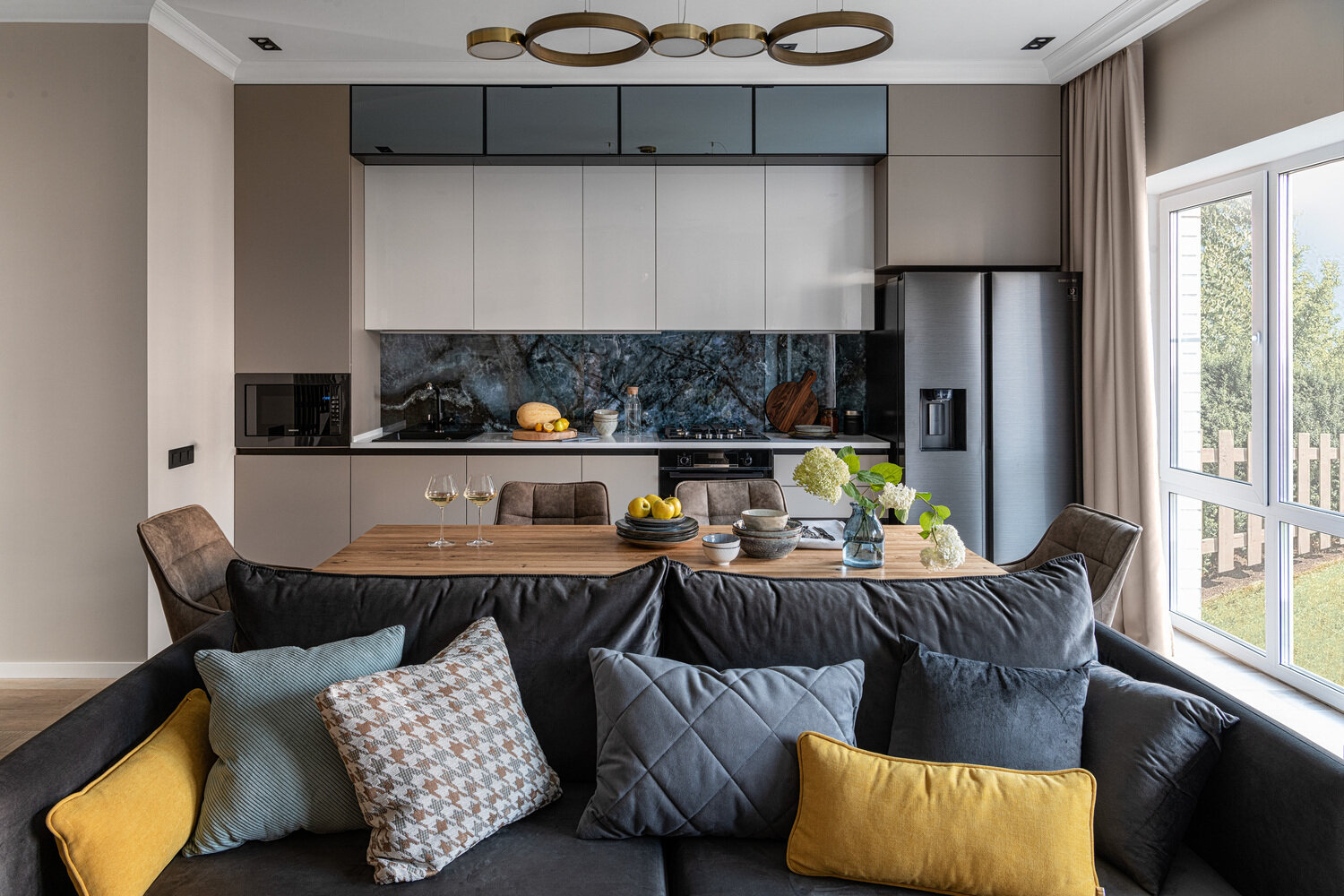 Отсутствие тюля на окнах в кухне-гостиной — пожелание заказчиков. Переехав в таунхаус с небольшим земельным участком, они решили, что теперь вид за окном на их мини-сад через большие окна в пол будет радовать их глаз в любое время года, и тюль или вуаль на окнах не нужны.