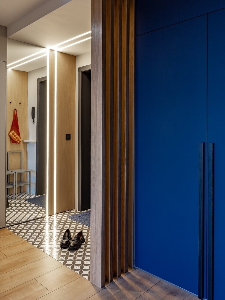 Прихожая создает общее впечатление о квартире. Первое, что привлекает глаз при входе — графичные линии освещения, отражения которых «играют» в зеркале. Шпонированные рейки поддерживают акцент и динамику вертикальных линий, разграничивая пространство входной зоны от основного коридора.